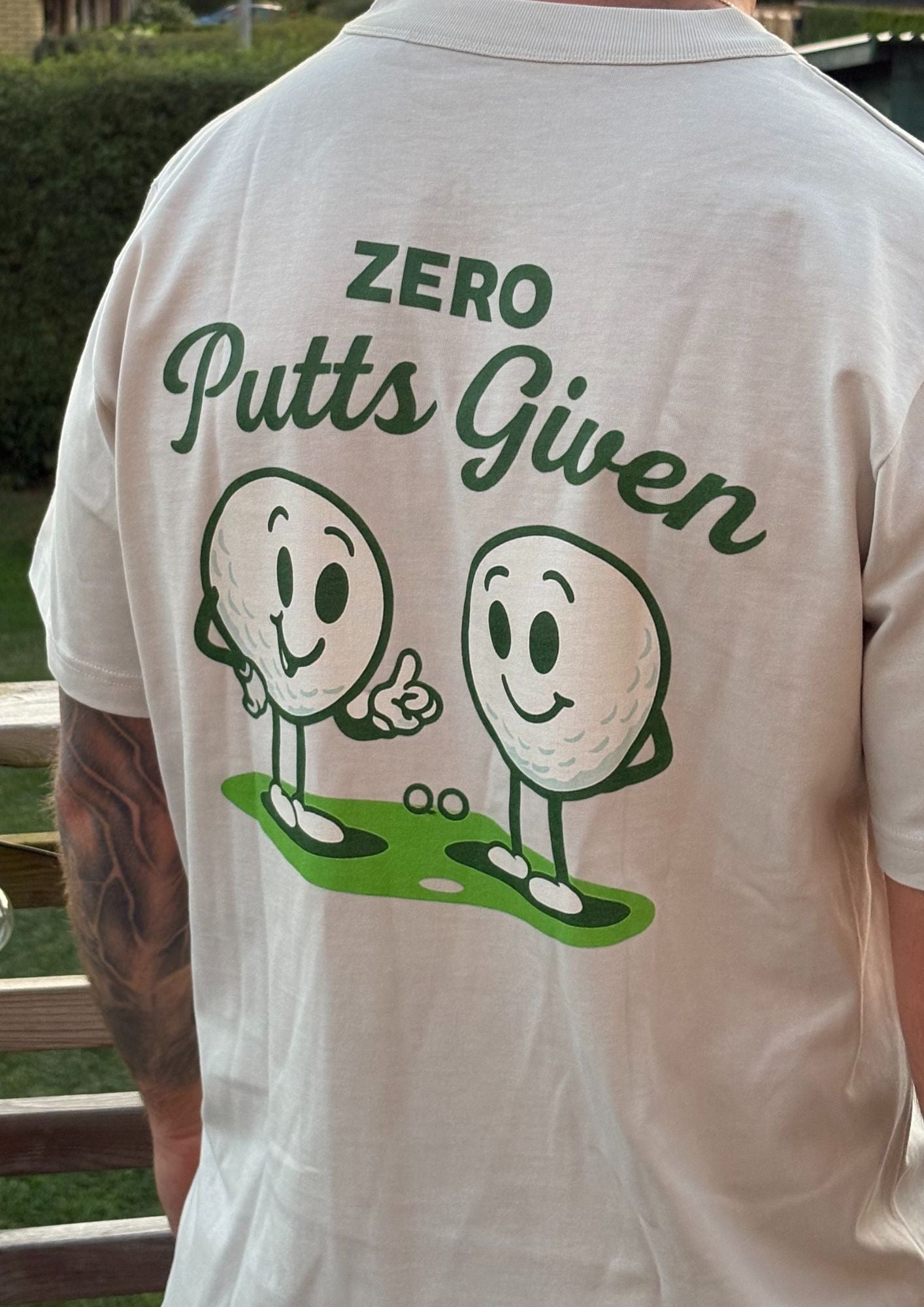 White shirt with 'Zero Putts Given' text and cartoon golf balls on the back, outdoors.